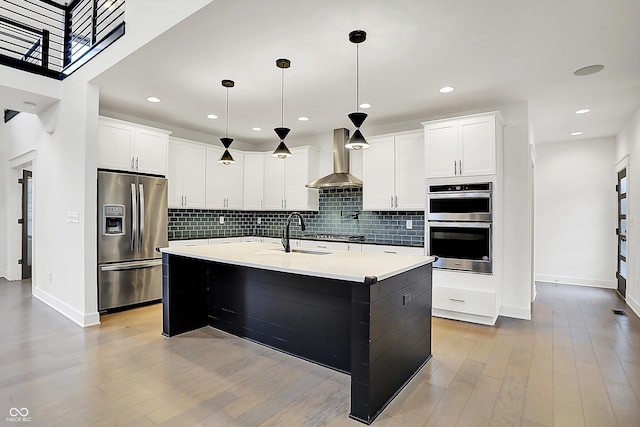 kitchen with appliances with stainless steel finishes, pendant lighting, white cabinets, a kitchen island with sink, and wall chimney exhaust hood