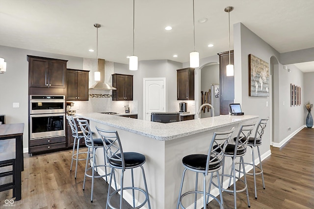 kitchen with double oven, wall chimney range hood, decorative light fixtures, and backsplash