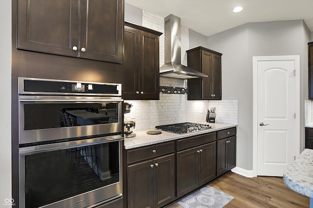 kitchen featuring dark brown cabinetry, light stone countertops, wall chimney exhaust hood, and appliances with stainless steel finishes