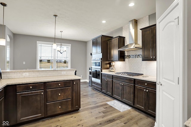 kitchen with dark brown cabinetry, light hardwood / wood-style flooring, pendant lighting, stainless steel appliances, and wall chimney range hood