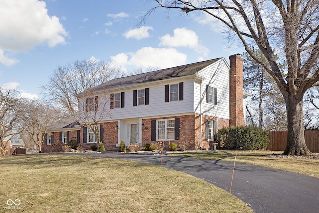 colonial-style house featuring brick siding, fence, driveway, a front lawn, and a chimney