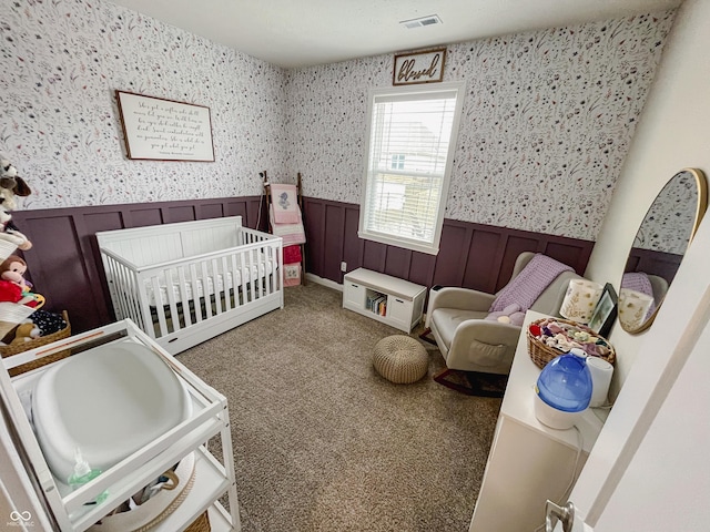 carpeted bedroom with a nursery area, wainscoting, visible vents, and wallpapered walls