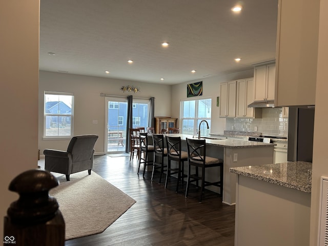 kitchen with dark wood-type flooring, tasteful backsplash, light stone countertops, an island with sink, and a kitchen bar