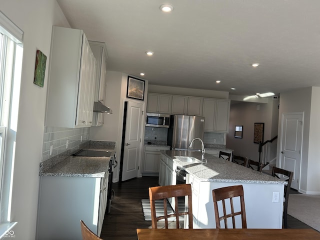 kitchen with light stone counters, appliances with stainless steel finishes, dark wood-type flooring, and a kitchen island with sink