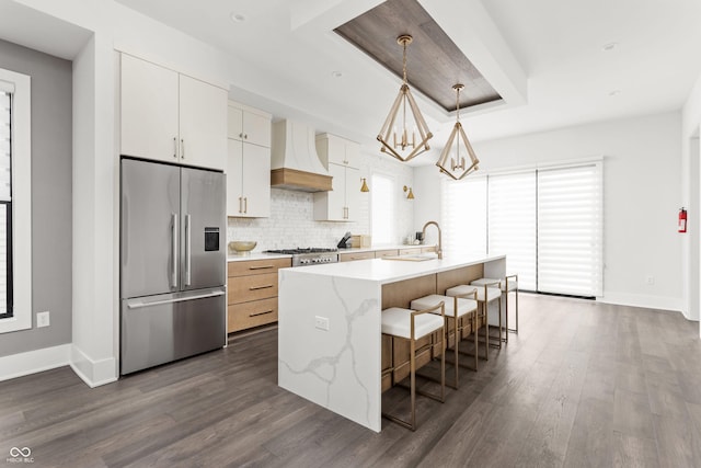 kitchen with white cabinetry, custom exhaust hood, hanging light fixtures, a kitchen island with sink, and stainless steel appliances