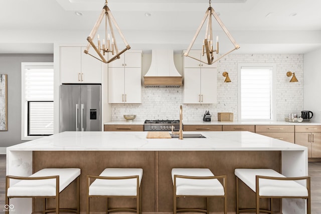 kitchen with white cabinetry, stainless steel fridge, a chandelier, custom range hood, and a center island with sink