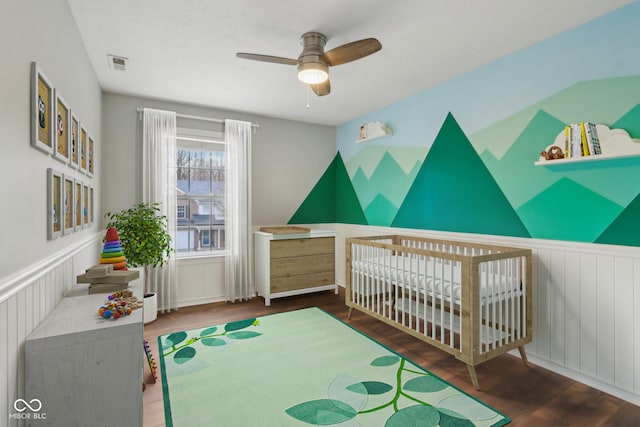 bedroom featuring visible vents, a nursery area, wood finished floors, and wainscoting