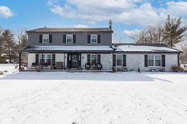 snow covered rear of property with covered porch, brick siding, and a chimney