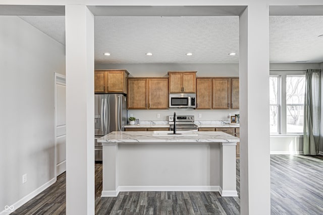 kitchen with appliances with stainless steel finishes, a kitchen island with sink, light stone countertops, dark wood-type flooring, and a textured ceiling