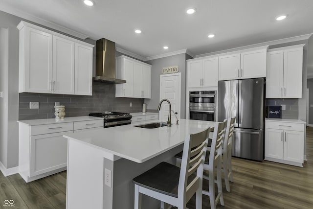 kitchen with wall chimney range hood, sink, white cabinetry, stainless steel appliances, and a center island with sink