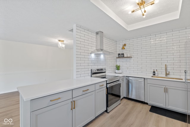 kitchen with light wood-style flooring, a sink, appliances with stainless steel finishes, wall chimney range hood, and a raised ceiling