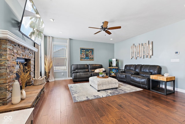 living room featuring a fireplace with raised hearth, ceiling fan, baseboards, and dark wood finished floors