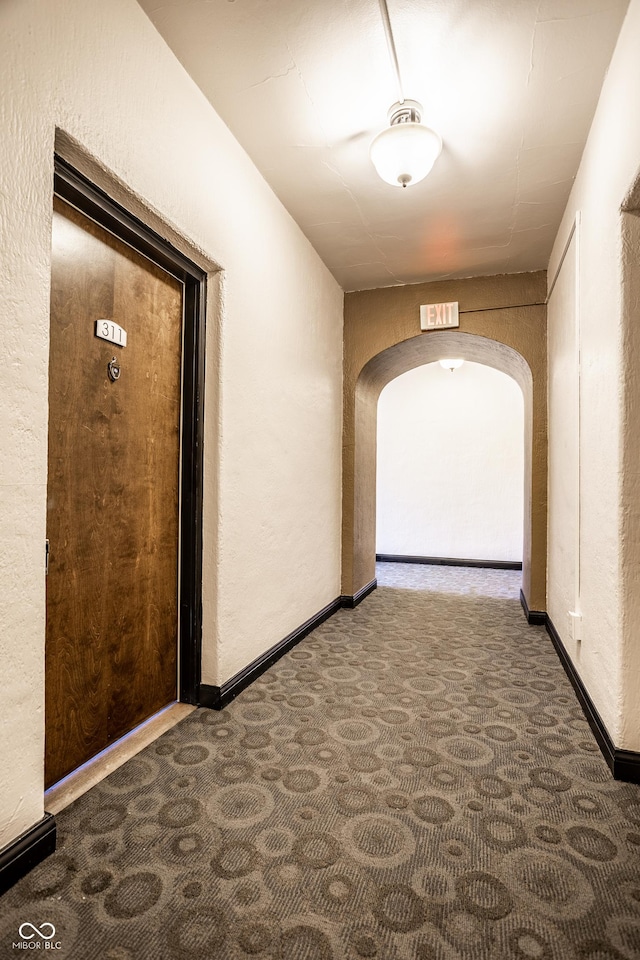 hall featuring baseboards, arched walkways, dark colored carpet, and a textured wall