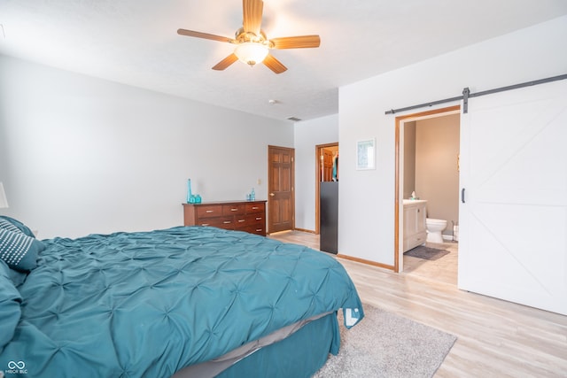 bedroom with a barn door, baseboards, ensuite bath, light wood-style flooring, and ceiling fan