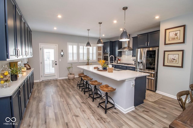 kitchen featuring an island with sink, decorative light fixtures, appliances with stainless steel finishes, wall chimney range hood, and a breakfast bar area