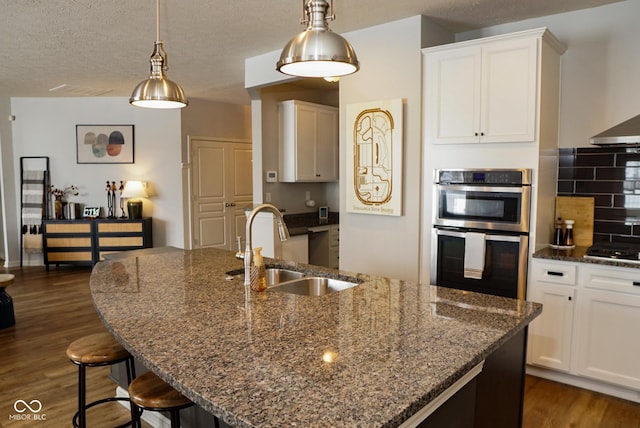 kitchen with double oven, a kitchen island with sink, white cabinets, and a sink