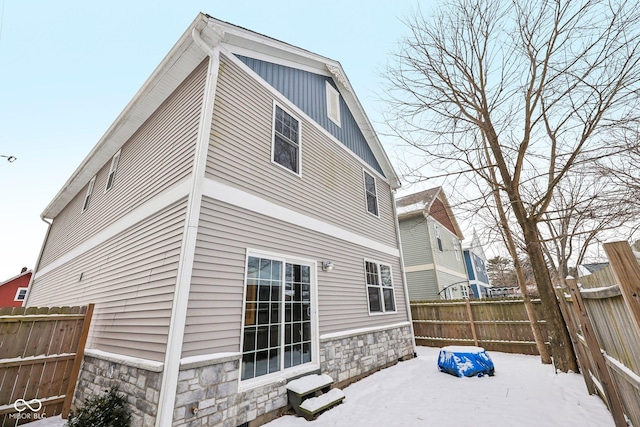 snow covered house featuring entry steps, stone siding, and fence