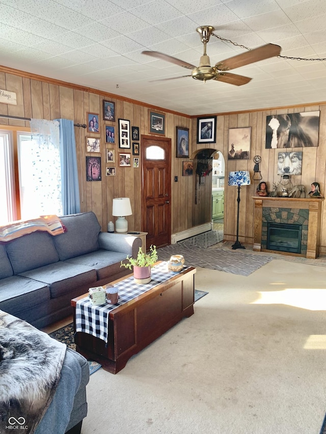 carpeted living room featuring a baseboard heating unit, wood walls, ceiling fan, crown molding, and a fireplace