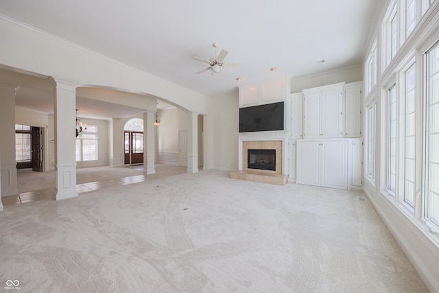 unfurnished living room featuring light colored carpet, ornamental molding, a tile fireplace, ornate columns, and ceiling fan with notable chandelier