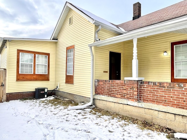 view of snow covered exterior with central AC unit, a chimney, and a shingled roof