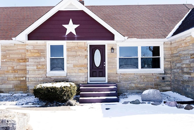 snow covered property entrance with stone siding and roof with shingles