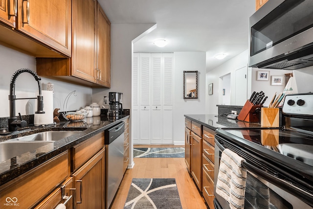 kitchen featuring light wood finished floors, brown cabinetry, dark stone countertops, stainless steel appliances, and a sink