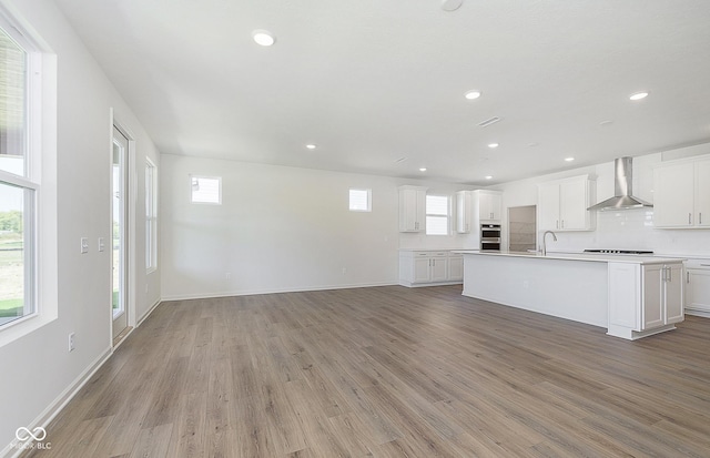 kitchen with a center island with sink, tasteful backsplash, light wood-style floors, open floor plan, and wall chimney range hood