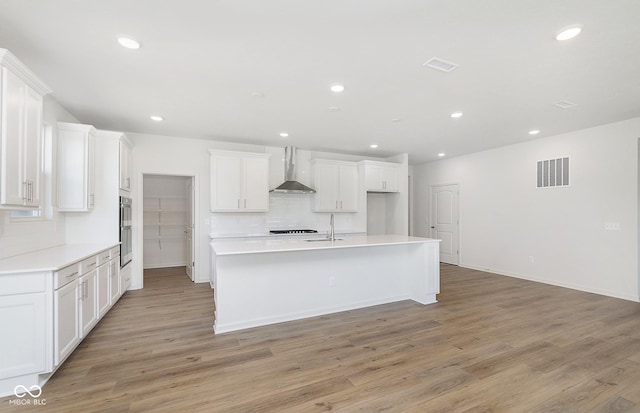 kitchen with visible vents, backsplash, light wood-style floors, a kitchen island with sink, and wall chimney exhaust hood