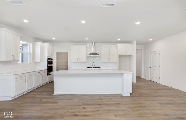 kitchen featuring visible vents, wall chimney exhaust hood, light wood-style flooring, stainless steel double oven, and white cabinetry