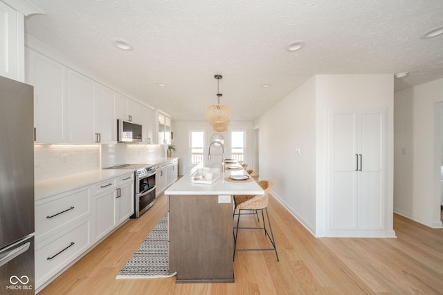 kitchen with decorative light fixtures, light countertops, appliances with stainless steel finishes, a kitchen island with sink, and white cabinetry