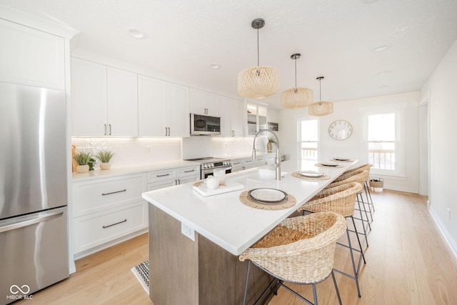 kitchen featuring a kitchen island with sink, stainless steel appliances, white cabinets, light countertops, and decorative light fixtures