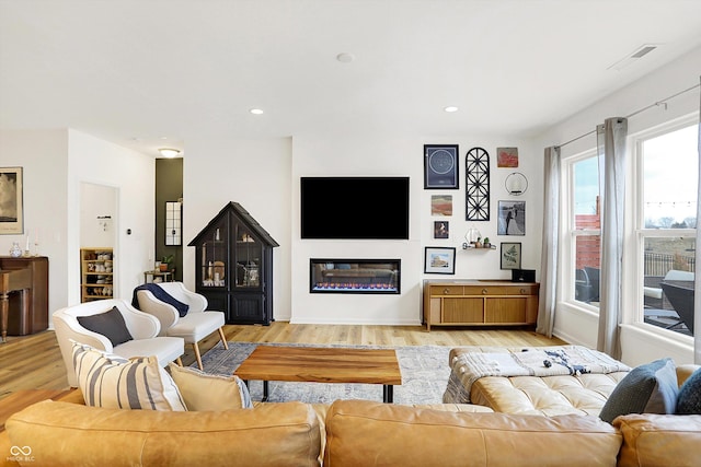 living room featuring light wood-type flooring, a glass covered fireplace, visible vents, and recessed lighting