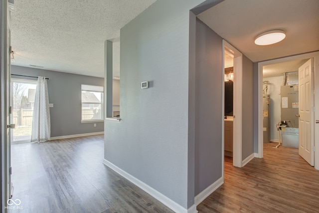 corridor featuring a textured ceiling, electric water heater, baseboards, and wood finished floors