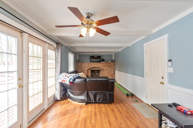 living room featuring ceiling fan, light wood-style flooring, a wainscoted wall, a fireplace, and beam ceiling