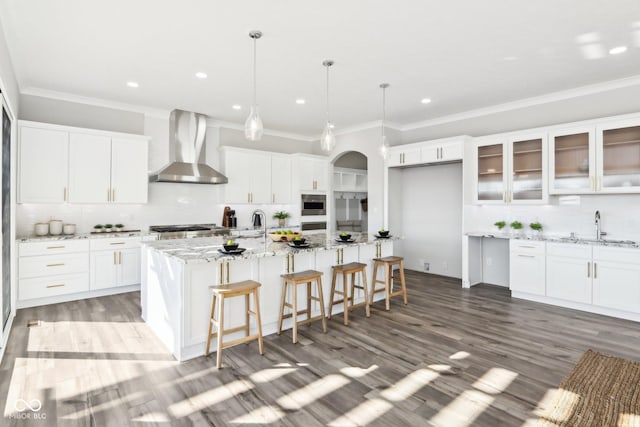 kitchen with an island with sink, wall chimney exhaust hood, glass insert cabinets, hanging light fixtures, and white cabinetry