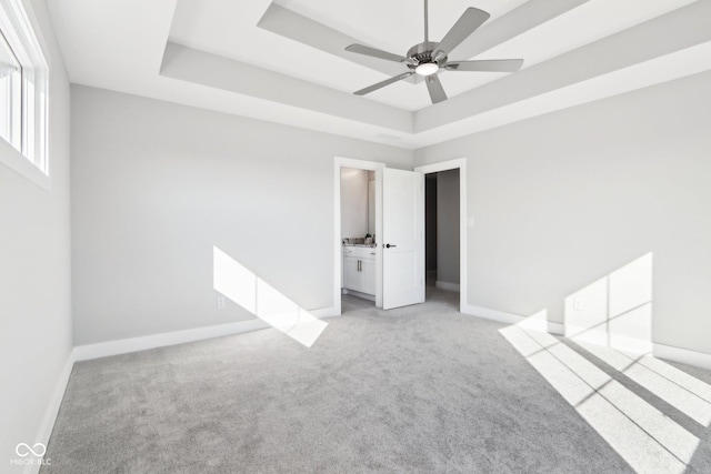 unfurnished bedroom featuring baseboards, a raised ceiling, light colored carpet, ensuite bath, and ceiling fan