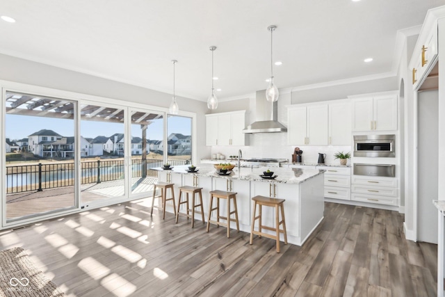 kitchen with light stone counters, white cabinets, wall chimney range hood, an island with sink, and decorative light fixtures