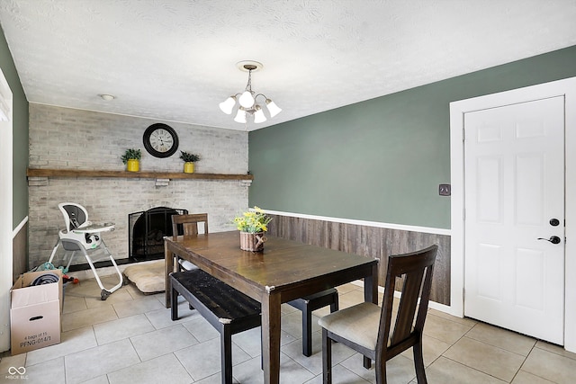 dining room featuring a textured ceiling, a chandelier, light tile patterned flooring, a wainscoted wall, and a brick fireplace