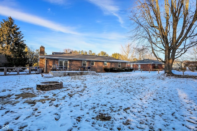 snow covered rear of property featuring brick siding, an outdoor fire pit, a chimney, and fence