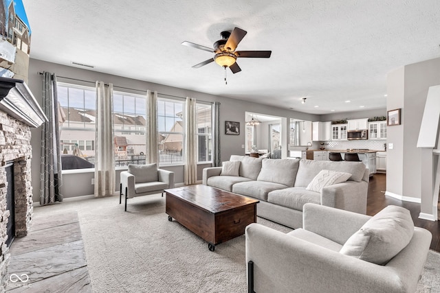 living area with visible vents, a ceiling fan, a stone fireplace, a textured ceiling, and baseboards