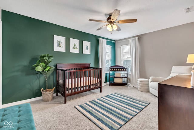 carpeted bedroom featuring a nursery area, baseboards, visible vents, and ceiling fan