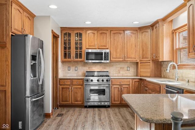 kitchen with brown cabinetry, appliances with stainless steel finishes, light stone counters, light wood-type flooring, and a sink