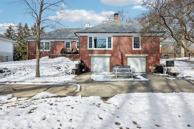 snow covered rear of property featuring driveway, a garage, a chimney, and brick siding