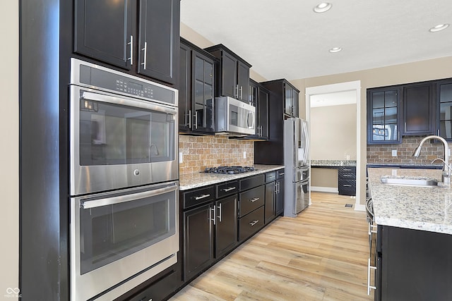 kitchen featuring light wood finished floors, glass insert cabinets, stainless steel appliances, dark cabinetry, and a sink