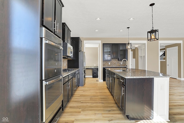 kitchen featuring stainless steel appliances, stone countertops, a sink, an island with sink, and light wood-type flooring