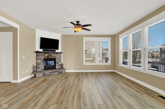 unfurnished living room featuring light wood-type flooring, ceiling fan, a stone fireplace, and baseboards