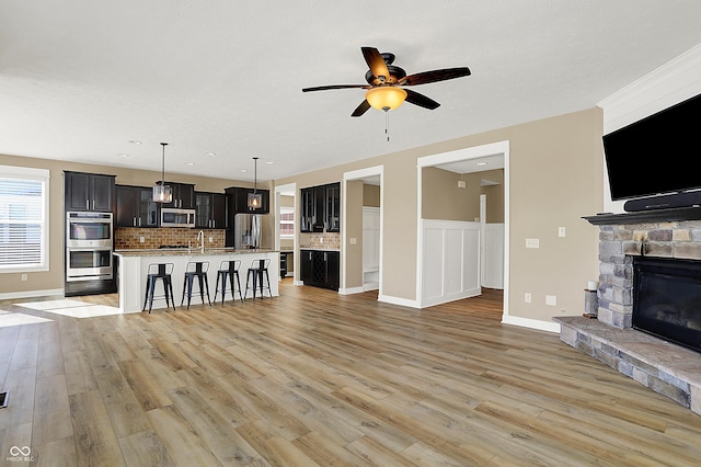 unfurnished living room featuring a wainscoted wall, light wood finished floors, a ceiling fan, a sink, and a stone fireplace