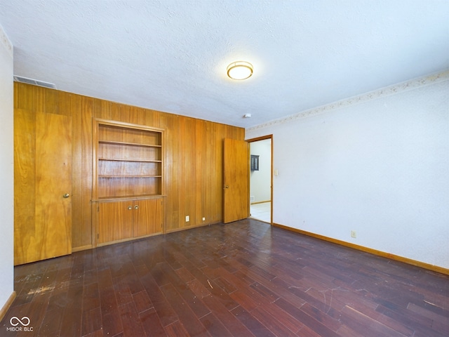 unfurnished room featuring dark wood-style floors, built in features, wood walls, a textured ceiling, and baseboards