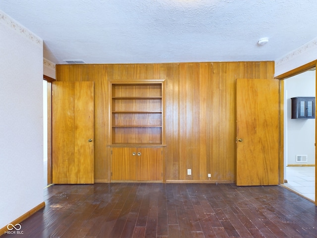 spare room with a textured ceiling, wooden walls, visible vents, built in features, and dark wood finished floors