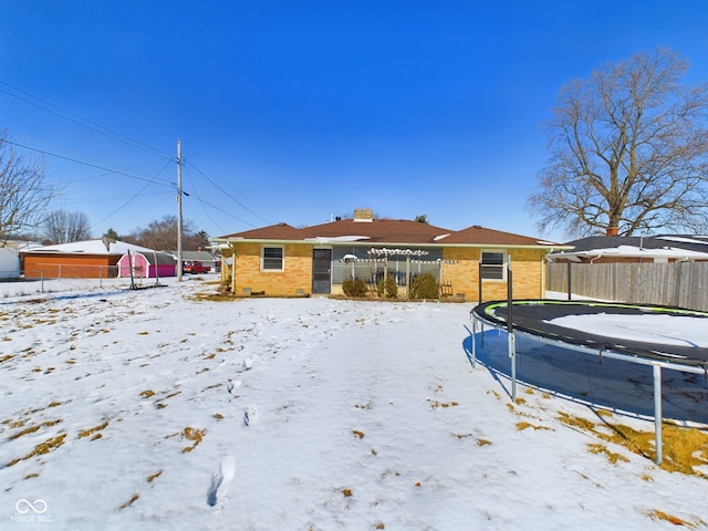 snow covered back of property featuring brick siding, a trampoline, and fence